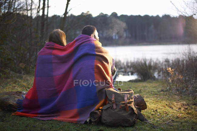 Jeune couple au bord du lac enveloppé dans une couverture au coucher du soleil — Photo de stock