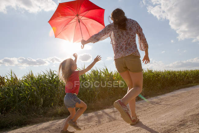 Mother and daughter walking through field carrying red umbrella — Stock Photo