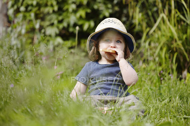 Little girl in hat sitting in grass eating snack — Stock Photo