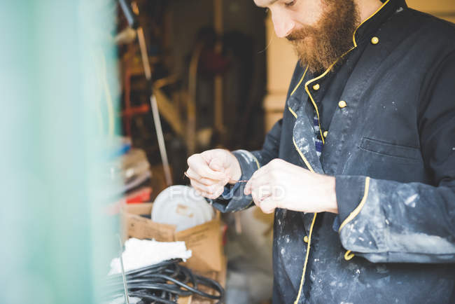Hombre barbudo en taller sosteniendo alambre mirando hacia abajo - foto de stock