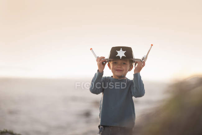 Boy dressed as cowboy sheriff holding onto hat and toy guns in sand dunes — Stock Photo