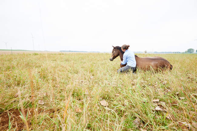 Woman crouching with arm around horse lying down in field — Stock Photo