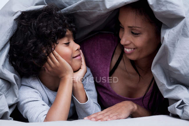 Madre e hijo escondidos debajo del edredón en la cama - foto de stock