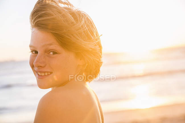 Retrato de niño en la playa mirando por encima del hombro a la cámara sonriendo - foto de stock
