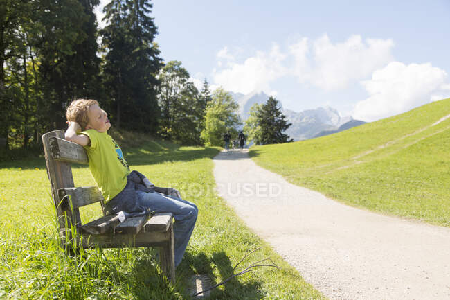 Niño sentado en el banco del parque en la carretera rural, Eckbauer bei Garmisch, Baviera, Alemania - foto de stock