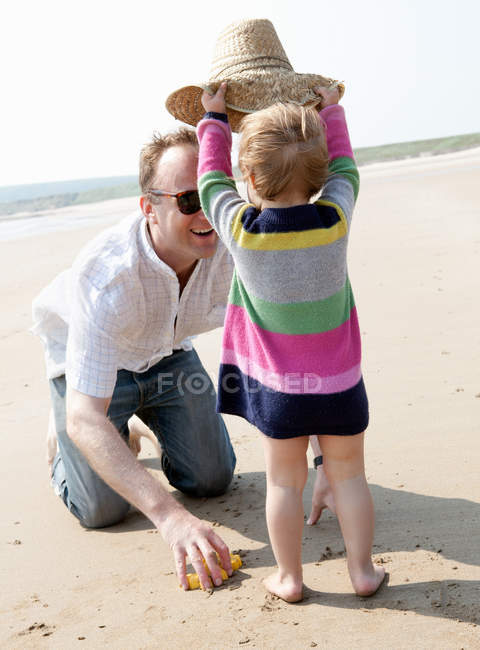 Chica poniendo sombrero en el padre en la playa - foto de stock