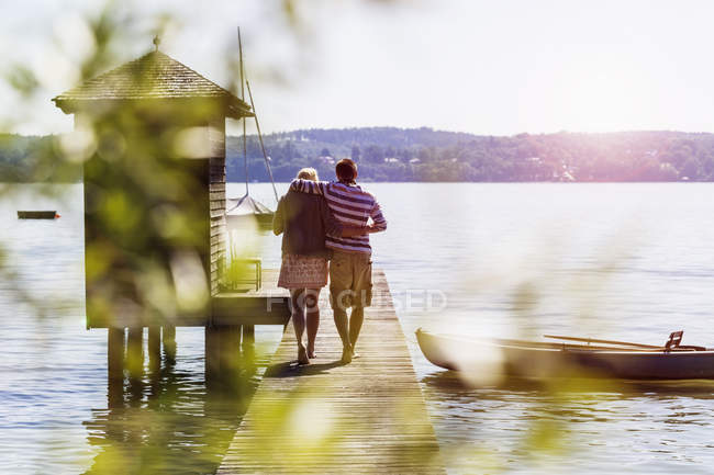 Rear view of young couple walking along pier on lake to boathouse, Schondorf, Ammersee, Bavaria, Germany — Stock Photo