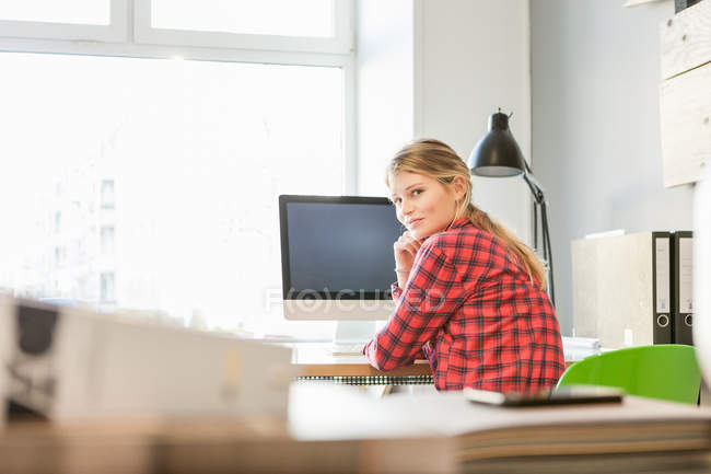 Vue arrière de la jeune femme à l'ordinateur regardant par-dessus l'épaule à la caméra souriant — Photo de stock