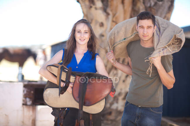 Young couple carrying saddle and sack of hay — Stock Photo
