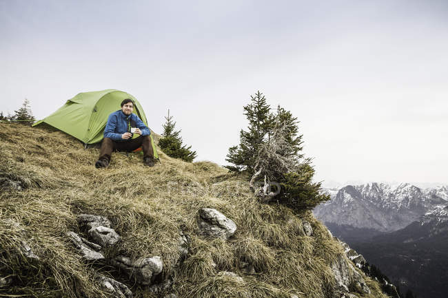 Young male hiker drinking coffee in front of tent on Klammspitze mountain, Oberammergau, Bavaria, Germany — Stock Photo