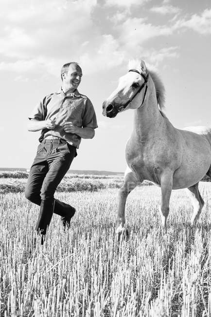 B&W image of male trainer running in front of white horse in field — Stock Photo