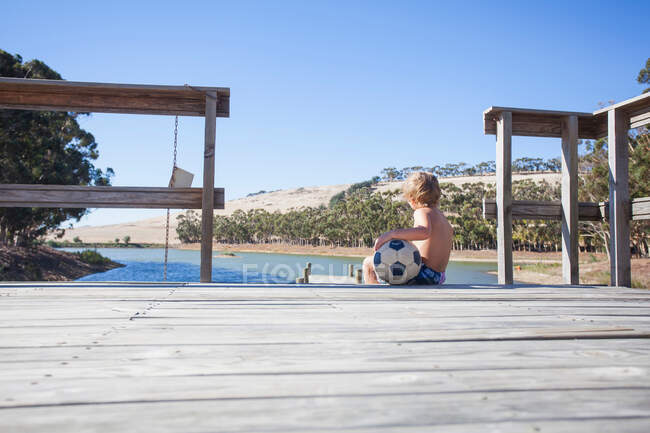 Junge sitzt mit Fußball auf Seebrücke — Stockfoto