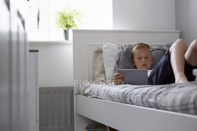 Blick durch die Tür eines Jungen, der auf dem Bett liegt und auf sein digitales Tablet blickt — Stockfoto