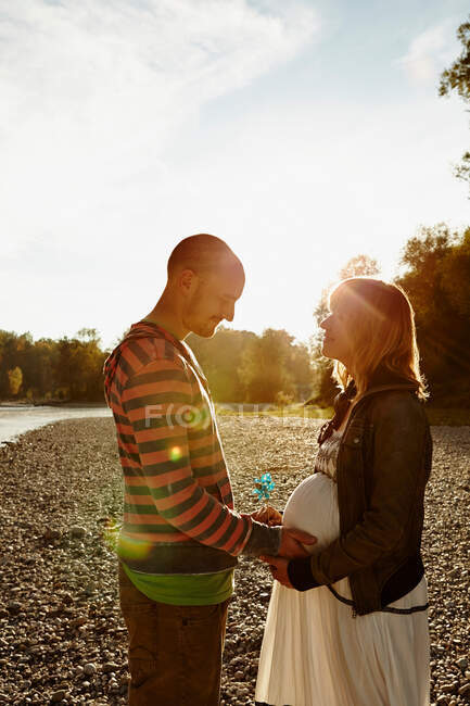Mujer embarazada y pareja de pie cara a cara junto al río sosteniendo molino de viento - foto de stock