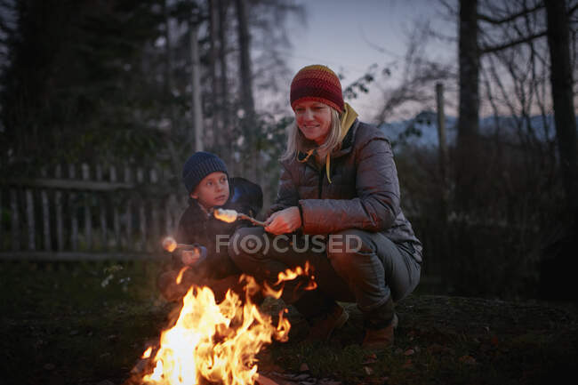 Mature woman and son toasting marshmallows on garden campfire at dusk — Stock Photo