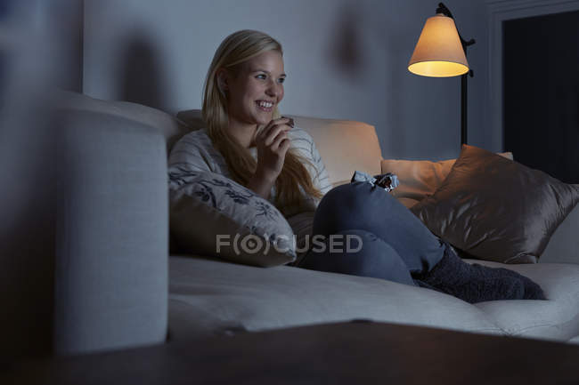 Jovem mulher sentada no sofá, assistindo tv, comendo chocolate — Fotografia de Stock