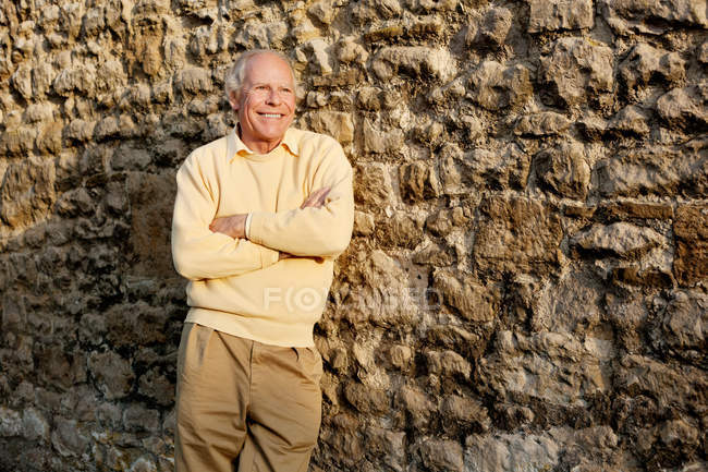 Hombre mayor apoyado en la pared de piedra con los brazos cruzados - foto de stock