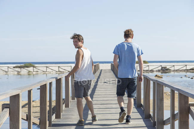 Full length rear view of young men walking on wooden pier, looking over shoulder, Sardinia, Italy — Stock Photo
