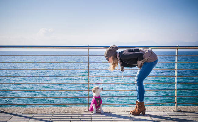 Full length side view of young woman by railings in front of ocean bending over looking at dog — Stock Photo