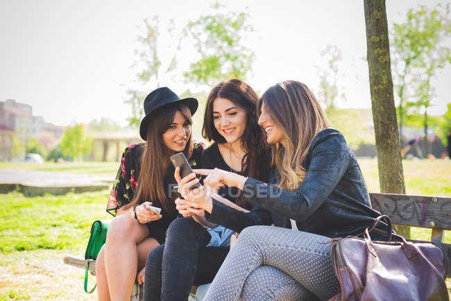 Trois jeunes amies utilisant un smartphone sur le banc du parc — Photo de stock