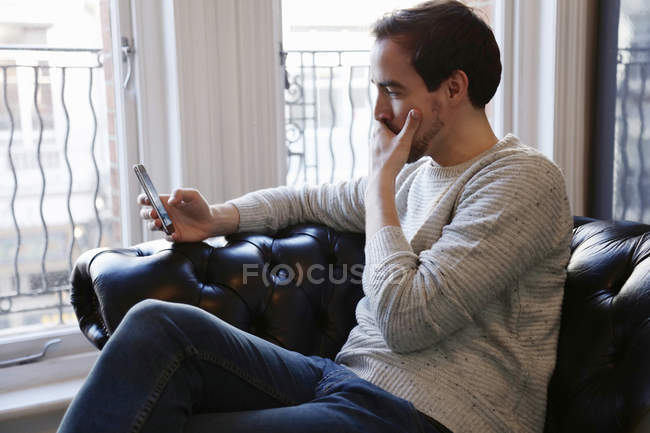 Homem adulto médio sentado no sofá, olhando para o telefone inteligente — Fotografia de Stock