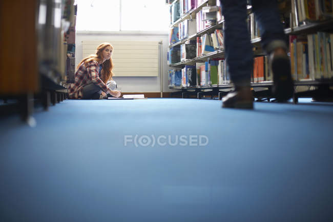 Joven estudiante universitaria mirando hacia arriba mientras trabaja en el piso de la biblioteca - foto de stock