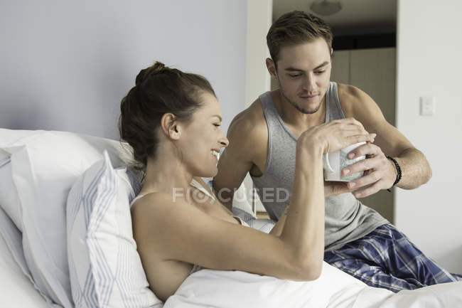 Young man handing coffee to girlfriend lying in bed — Stock Photo