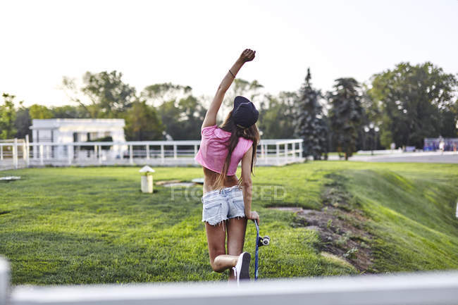 Young woman walking through park, carrying skateboard, punching air, rear view — Stock Photo
