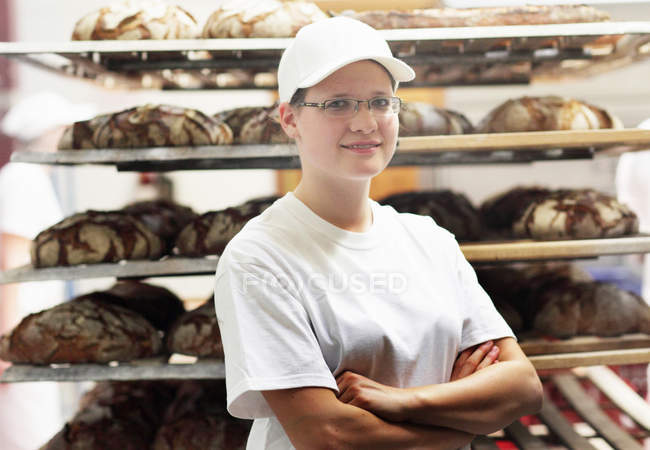 Padeiro em pé na frente de pão em prateleiras com os braços dobrados — Fotografia de Stock