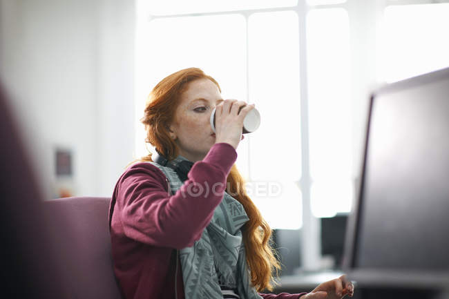 Молода студентка коледжу за комп'ютерним столом п'є каву — стокове фото