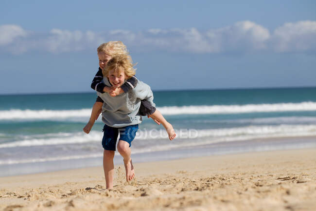 Мальчик несет брата на спине по пляжу — стоковое фото