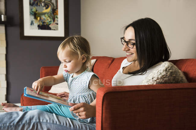 Mother teaching daughter to read book on sofa at home — Stock Photo