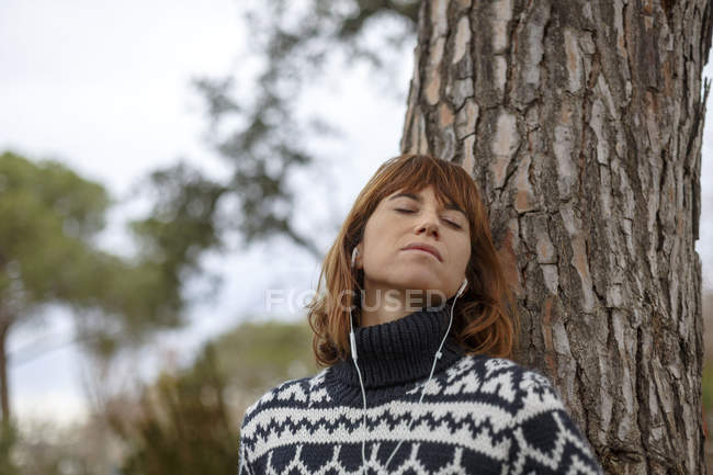 Mulher encostada à árvore usando fones de ouvido, olhos fechados — Fotografia de Stock