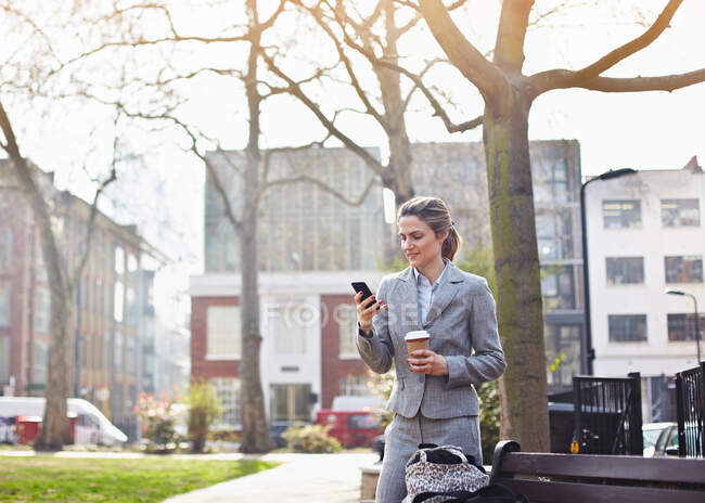 Бізнес-леді, використовуючи мобільний телефон в парку з кавою — стокове фото