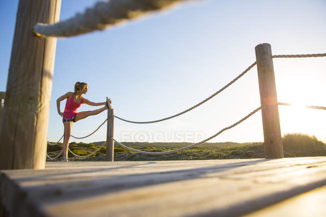 Young woman on wooden pathway, exercising, stretching leg, low angle view — Stock Photo
