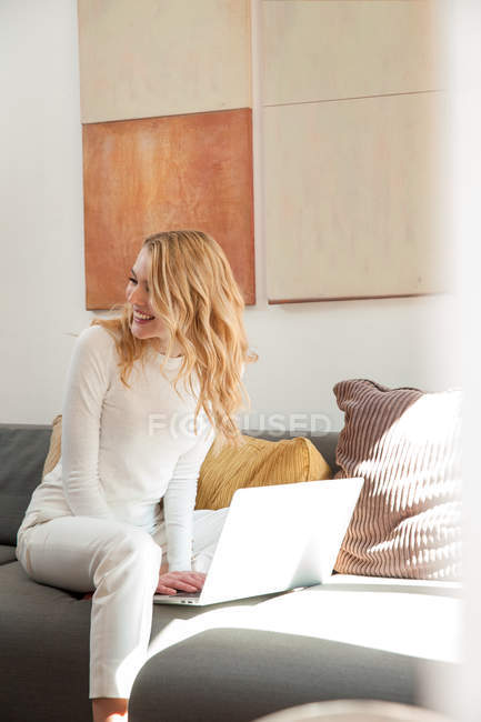 Женщина на диване с помощью ноутбука смотрит в сторону улыбаясь — стоковое фото