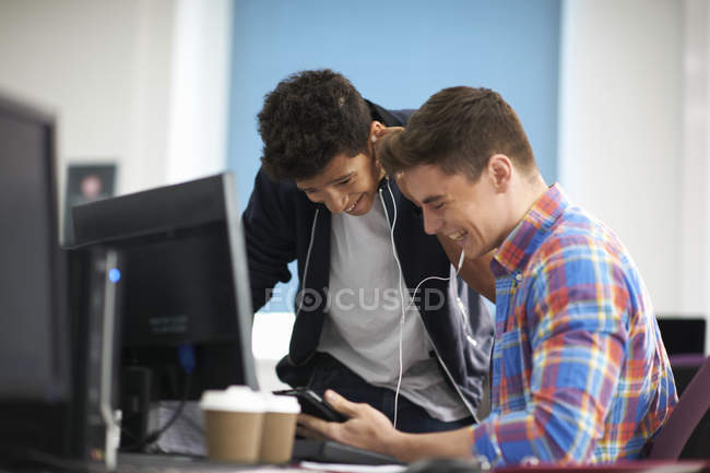 Jóvenes estudiantes universitarios en el escritorio de la computadora riendo mientras escuchan los auriculares - foto de stock