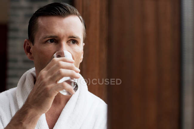 Человек в халате пьет воду у окна — стоковое фото