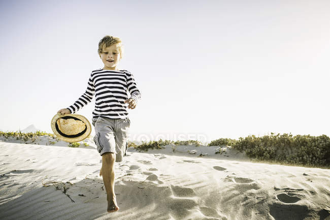 Молодий хлопець біжить уздовж пляжу, тримаючи солом'яний капелюх — стокове фото