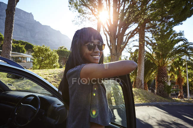 Portrait of young woman standing beside car, leaning on open car door — Stock Photo