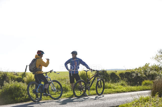 Ciclistas sosteniendo bicicletas en carretera rural charlando - foto de stock