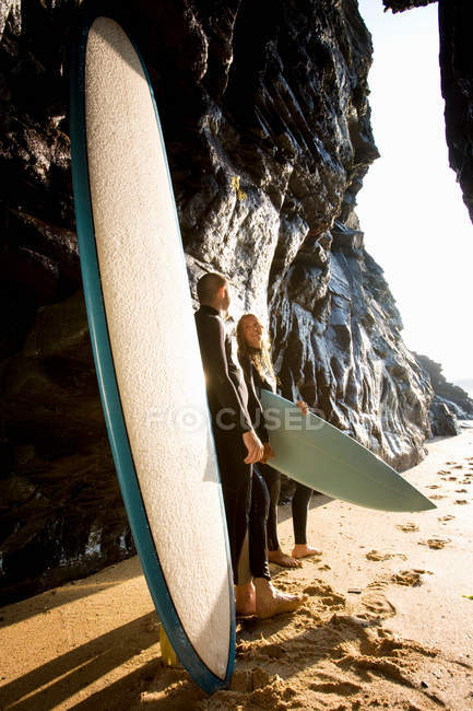 Pareja de pie con tablas de surf por grandes rocas sonriendo . - foto de stock