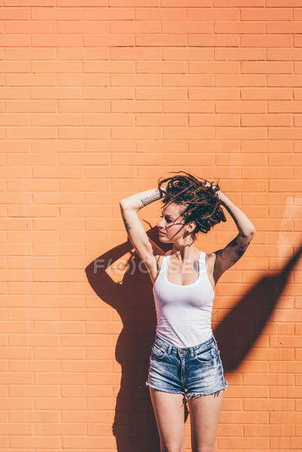 Jovem mulher com as mãos no cabelo dreadlock na frente da parede laranja — Fotografia de Stock