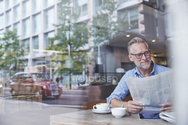 Mature man sitting in cafe, reading newspaper, street reflected in window — Stock Photo