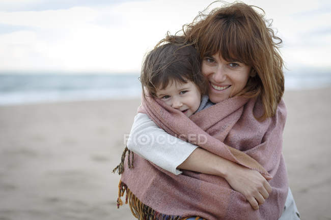 Madre e hija envueltas en manta abrazándose en la playa - foto de stock