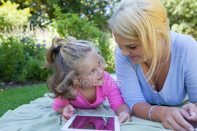 Madre e hija tumbadas en una manta en el jardín usando una tableta digital - foto de stock