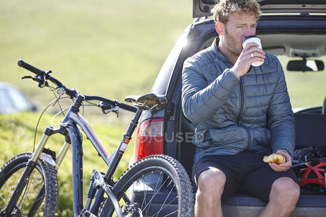 Ciclista sentado na bota do carro beber de copo descartável — Fotografia de Stock