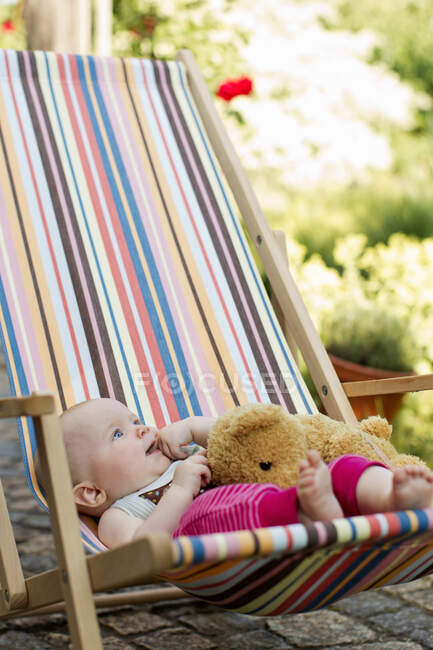 Baby girl sitting on deck chair with teddy bear — Stock Photo