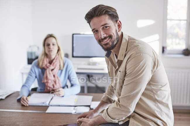 Чоловік сидить на столі в офісі, дивлячись на камеру посміхаючись — стокове фото
