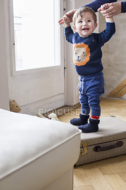 Baby boy holding mothers hands and standing on suitcase — Stock Photo